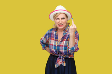 Portrait of angry modern stylish mature woman in casual style with hat and eyeglasses standing with middle finger fuck sign and looking at camera. indoor studio shot isolated on yellow background.