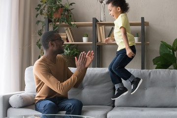 African father clapping hands looking at son jumping on sofa