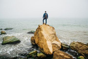 Traveler with a backpack stands on a rock against a beautiful sea with waves, a stylish hipster boy posing near a calm ocean during a wonderful journey around the world. Shoot from the back