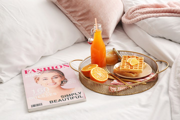 Tray with tasty breakfast and fashion magazine on bed