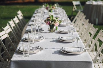 A large white long table, decorated with fresh flowers, with plates, glasses and forks, stands in a park with green grass. Wedding decorations and details. Preparing for a wedding party.
