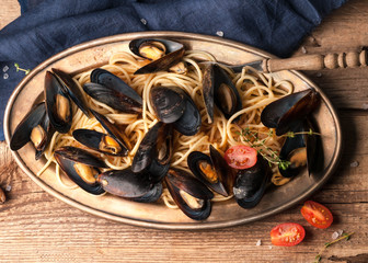 top view of delicious mussels in shells cooked in sauce with pasta and red tomatoes on golden metal plate with blue cloth on wooden background