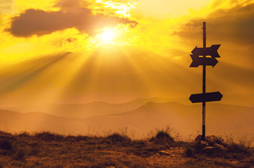 Blank signpost arrows ponting in opposite directions with sun rays over the mountains in the background
