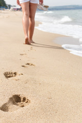 Young girl walking on sandy beach leaving footprints in the sand. Small sea waves. Beach summer travel concept