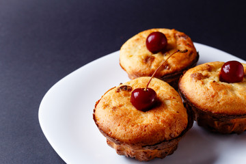 Three homemade cupcakes with cherry on the top on white plate with dark background. Horisontal image with copyspace left