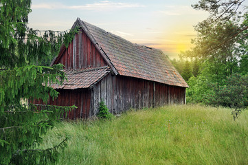 Obraz premium Overdue barn with a sunset in the horizon. Summer view during early night in Sweden. Pine and meadow in the foreground. Landscape with a calm feeling the west of Sweden.