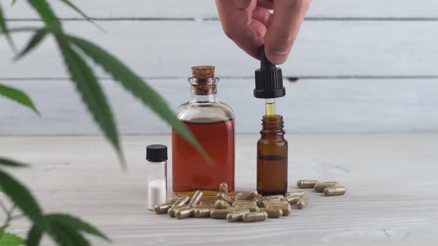 Hand dropping CBD oil from pipette to the bottle on White table with Marijuana plant CBD pills and CBD crystals