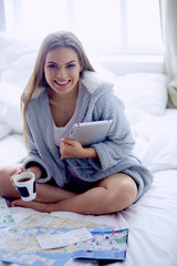 Relaxed young woman sitting on bed with a cup of coffee and digital tablet