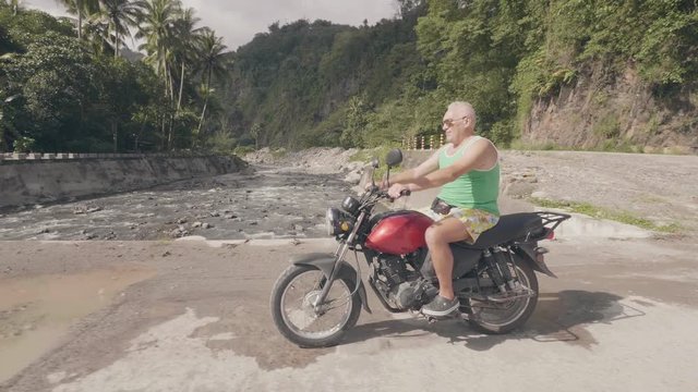 Elderly man driving on motorcycle on countryside road while moto trip at summer holiday. Traveler man riding on motorbike on river bridge in asian village. Moto travel.