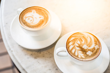 Two cups of coffee with beautiful leaf and pig latte art and sun flare lighting on a marble table in a coffee shop in the morning.