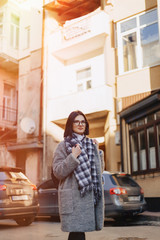 Attractive positive young girl wearing glasses in a coat on the background of buildings on cars