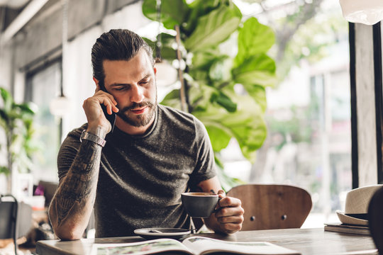 Handsome bearded hipster man use smartphone and reading book with coffee at table in cafe.Communication and technology concept