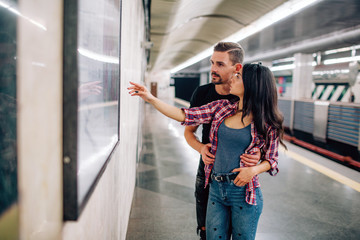 Fototapeta premium Young man and woman use underground. Couple in subway. Casual people in metropolitan. Young woman point on wall and smile. Guy hold hands on her hips.
