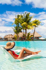 Luxury hotel swimming pool woman relaxing in lounging chair enjoying summer vacation. Tourist with hat and red swimsuit in water lounger.