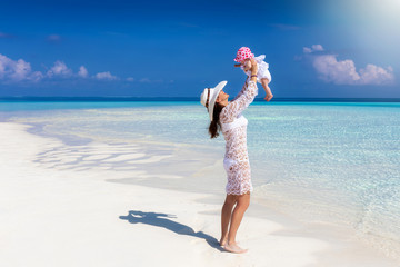 Glückliche Mutter spielt mit ihrem kleinen Baby am tropischen Strand mit türkisem Meer und blauem...