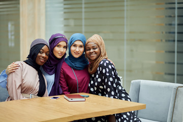 Portrait of smart elegantly dressed caucasian and african muslim female friends look at camera with cheerful expression at office background