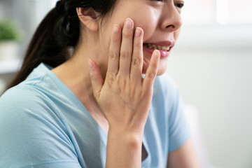 Young asian girl with toothache and dental problems. japanese casual woman hands touching face feeling sensitive teeth inside. frowning unhappy discomfort wife illness indoors in blue t shirt.