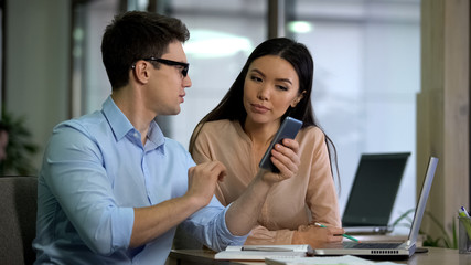 Male employee showing attractive asian colleague how to use dating app on phone
