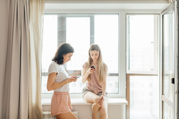 Two beautiful girls using their smart phones, standing in front of the window at home. close up photo.women prefer online comunication to real one