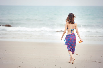 Fototapeta na wymiar Back view of shapely beautiful girl walking on tropical beach in swimsuit and holding orange juice in nature with blue sky