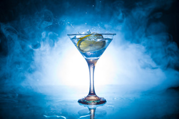 Martini cocktail glass in hand splashing on dark toned smoky background or colorful cocktail in glass with splashes and olives. Party club entertainment. Mixed light. Selective focus