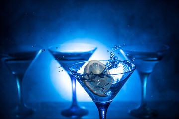 Martini cocktail glass in hand splashing on dark toned smoky background or colorful cocktail in glass with splashes and olives. Party club entertainment. Mixed light. Selective focus
