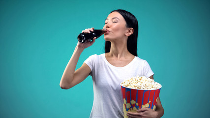 Asian woman holding big paper cup with popcorn and drinking soda visiting cinema
