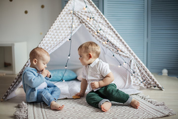 Two gorgeous infant boys of the same age sit on carpet in teepee tent, have fun and play with hristmas garlandin a cozy nursery.