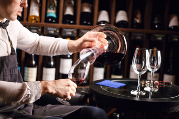 Wine taster or degustator mixing red wine into carafe to make perfect color on background with wine bottles racks in wine house.