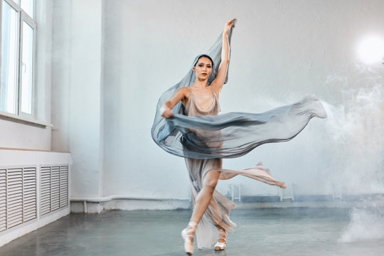 Full size portrait of talanted ballet student woman wears grey fluttering scenic dress giving a performance on a final exam in light studio on white smoky background.