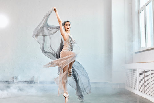Full-size expressive photo of female ballet dancer dressed in grey-blue flowing fabric spinning on one leg on white y background.