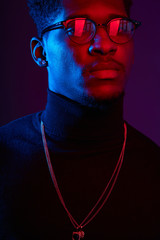 Obraz premium Low key studio shot of young handsome African man in glasses wearing smart poloneck with amulet on neck against dark violet background