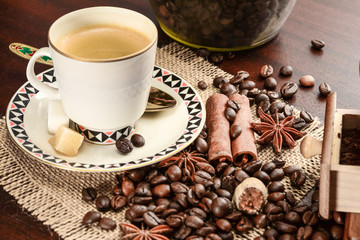 Cup of coffeee with saucer on a jute napkin. Vintage still life on wooden table with sugar, anise, cinnamon, grinder and coffee jar, close up