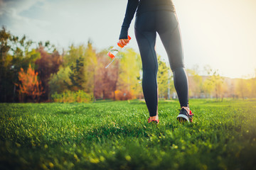 young girl feet in snickers standing in park and making running exercise on sunset and holding bottle of water in hand - fitness sport health concept