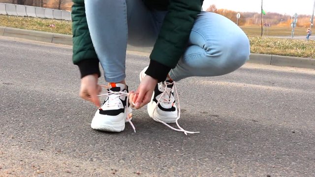 The girl untie the laces in white sneakers. Young Woman untying shoelaces of white sneaker standing on a road.