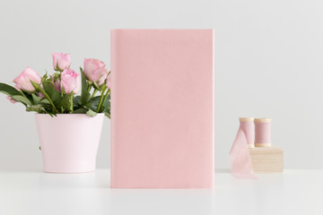 Pink book mockup with silk ribbons and pink roses in a pot on a white table.