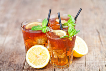 Ttree glasses of traditional iced tea with lemon, mint leaves and ice cubes on rustic wooden table.