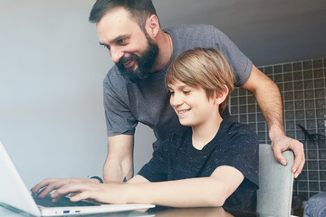 Handsome bearded young father in casual clothes and his cute little son using a laptop while at the table in the living room