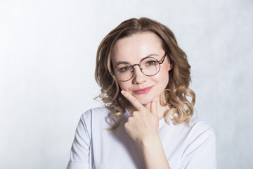 Fototapeta premium Smiling beautiful young woman with glasses holds her chin, looking around, tries to make a decision.