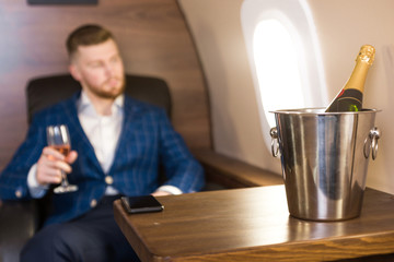 A young successful businessman in an expensive suit sits in the chair of a private jet with a glass of champagne in his hand and looks out the window