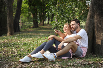Naklejka premium young caucasian couple people are in love, sitting and embracing and pointing at someone or something in park during summer season with trees