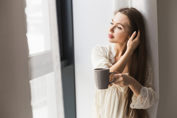 Attractive young girl in night clothes standing by the window with a mug of coffee
