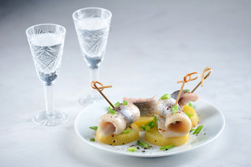 Slices of herring on boiled potatoes. Traditional Russian snack under vodka. Two glasses with vodka in the background. Close-up. Light background.