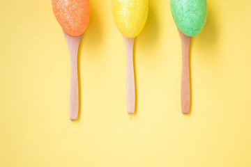 Three Colorful Easter eggs on pale yellow background, egg in wooden spoons, green, orange, yellow, egg hunt decoration, top view - flat lay