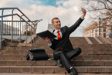 young man in a black suit and red tie takes a selfie on a smartphone. man with laptop sits on the steps in the city.