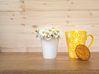 View of a bright yellow cup of coffee, a cookie and a bunch or bouquet of blooming daisies in a white porcelain vase on wooden table against a wooden background. Hello spring or good morning coffee  