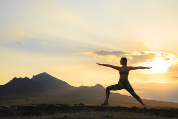 Silhouette of young woman practicing yoga or pilates at sunset or sunrise in beautiful mountain location.