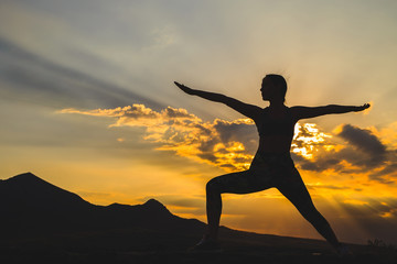 Fototapeta premium Silhouette of young woman practicing yoga or pilates at sunset or sunrise in beautiful mountain location.