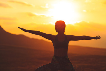 Silhouette of young woman practicing yoga or pilates at sunset or sunrise in beautiful mountain location.