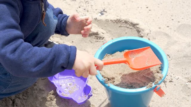 Closeup hands of little boy (baby, toddler),  child is playing with sand on a beach. Plastic kids pails buckets with shovels, molds. 